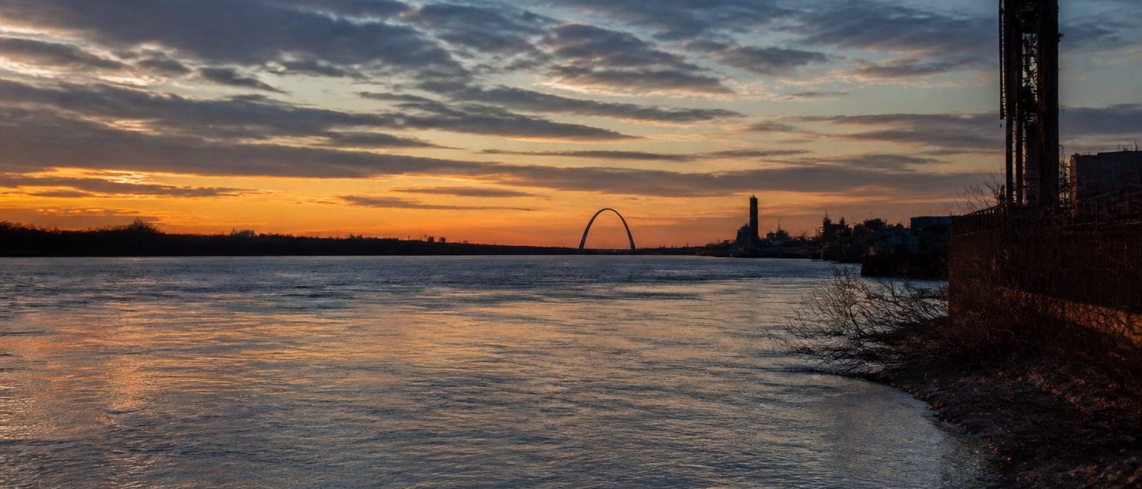 The Gateway Arch on the horizon over the Mississippi at dusk, seen from the riverbank