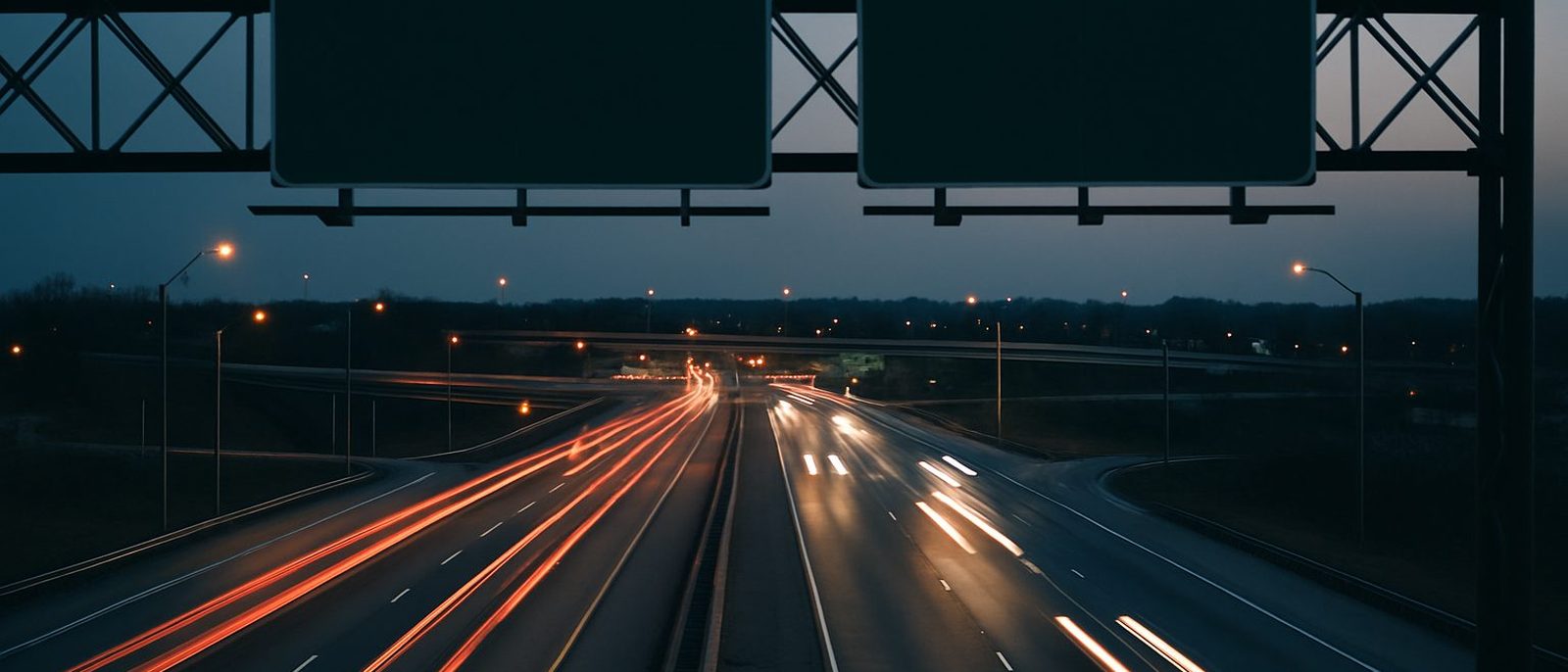 Six lanes of interstate traffic streaking under an overhead sign gantry at dusk in the St. Louis suburbs